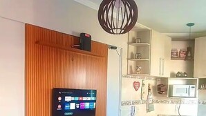 A modern living room with a wooden accent wall, a mounted flat-screen TV, a black ottoman, and a kitchen area with a microwave and cabinets. - Apto Boqueirão próximo ao Mar (Praia Grande)