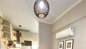 A living room with a brown sofa, a black ottoman, and a red throw blanket. There is a kitchen area with white cabinets and a window. - Apto Boqueirão próximo ao Mar (Praia Grande)