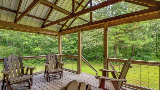 Eine überdachte Terrasse mit Adirondack-Stühlen und einem Tisch, mit Blick auf ein bewaldetes Gebiet.