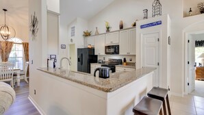 Una cocina moderna con armarios blancos, encimera de granito y electrodomésticos de acero inoxidable. Cuenta con un comedor con mesa y sillas blancas, y una araña colgante.