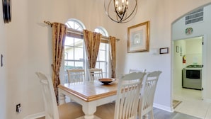 Un comedor con una mesa de madera y sillas blancas, ventanas amplias con cortinas y un candelabro.