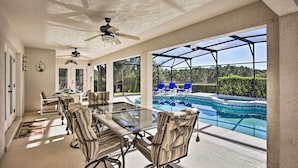 Un patio techado con mesa y sillas de vidrio, un ventilador de techo y vista a una piscina.