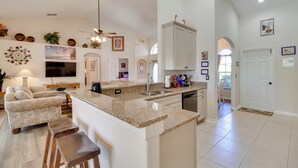 Una cocina amplia con una isla grande, un área de comedor con sofá y taburetes de bar, y una sala de estar con televisor y objetos decorativos.