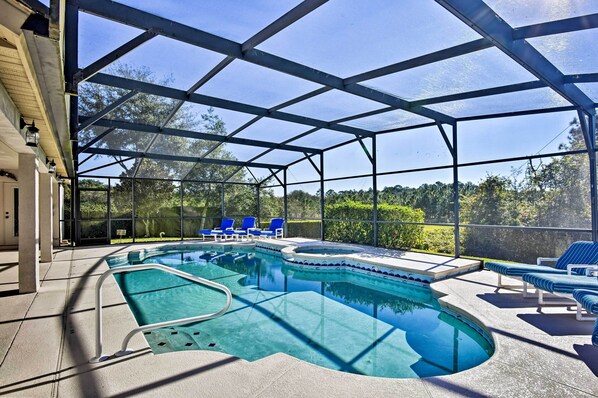 A covered swimming pool with blue chairs and a hot tub, surrounded by glass walls and trees.