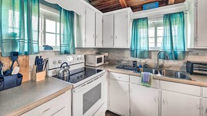 Cuina amb armaris blancs, una cuina, un forn microones i un rentaplat. Hi ha cortines color turquesa, un cartell blau amb números i una finestra.