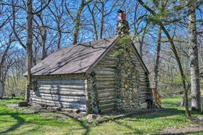 Une cabane rustique en rondins de bois, dotée d’une cheminée en pierre, entourée d’arbres.