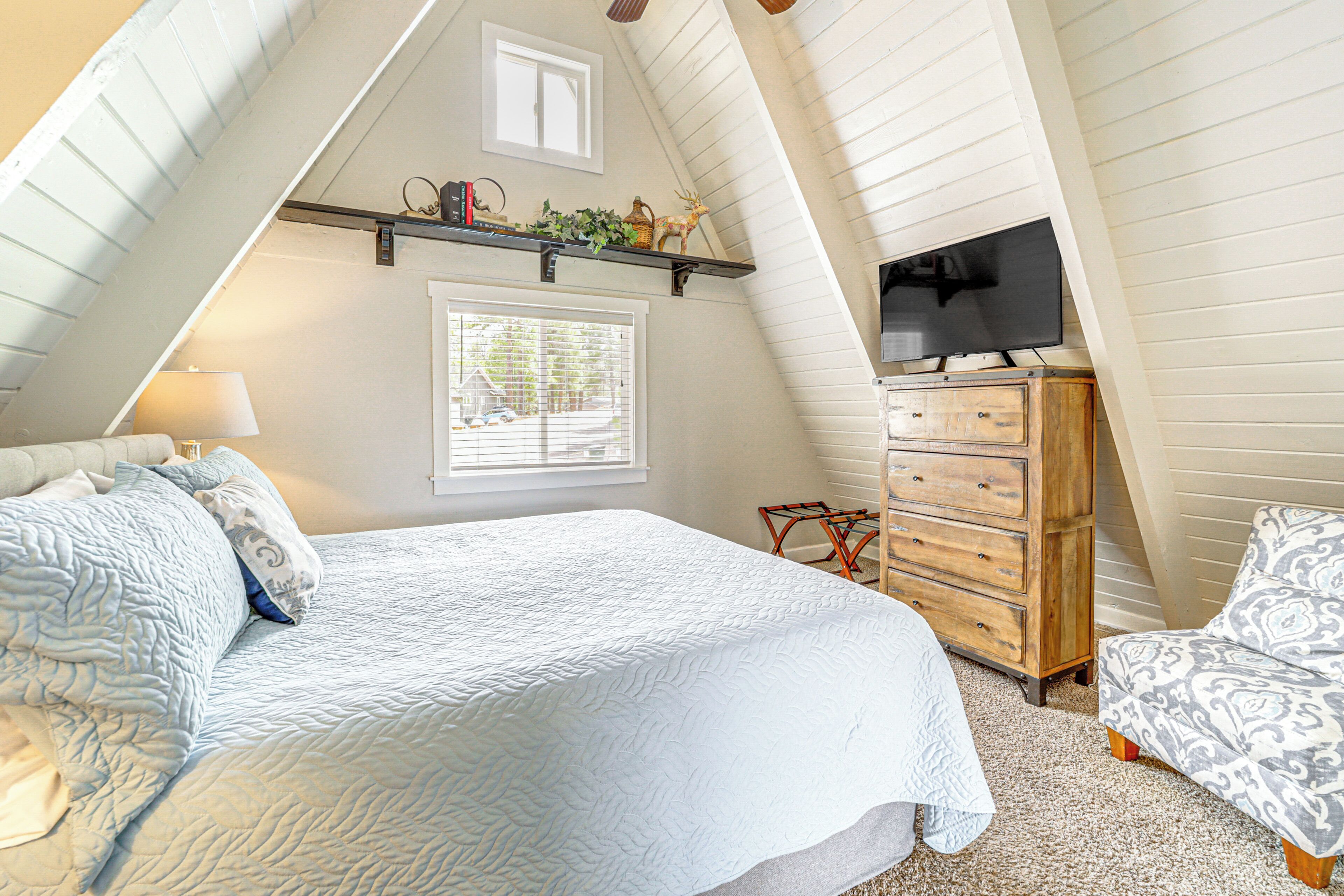 A bedroom with a sloped ceiling, a bed with a white quilt, a dresser, a television on a shelf, and a window with blinds.