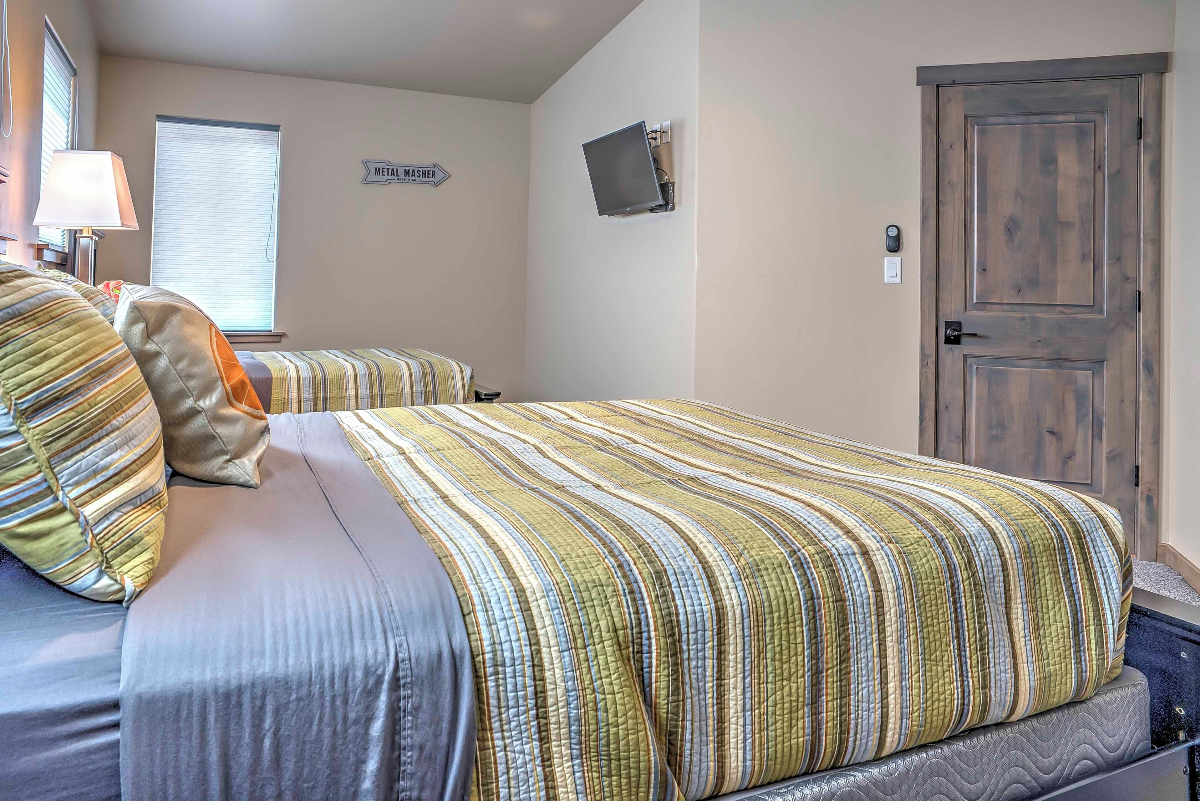 A room with two single beds, a window with blinds, a door, and a wall-mounted TV.