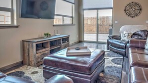 A living room with a large flat-screen TV, leather sofas, a wooden entertainment unit, and a large window with blinds. - Upscale Moab Home w/ Red Rock Views & Hot Tub (Moab)