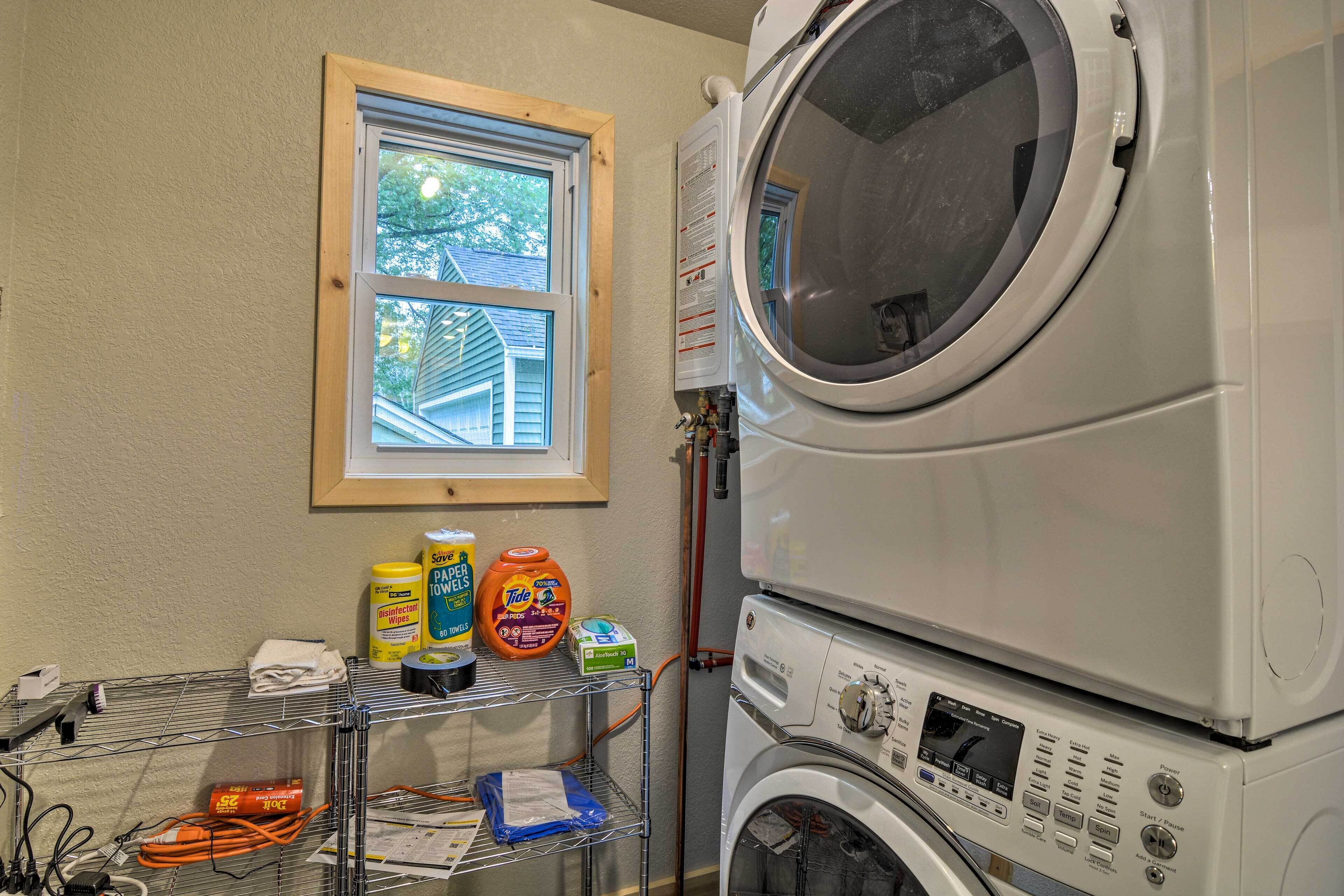 A laundry room with a stacked washer and dryer, a window with a view of a house, and shelves with cleaning supplies.