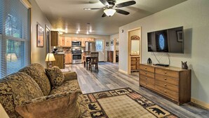 A living room with a sofa, a TV, a ceiling fan, and a kitchen in the background. - Pet-friendly Petoskey Area Cabin, Walk to River! (Alanson)
