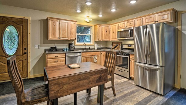 A kitchen with wooden cabinets, stainless steel appliances, and a dining area with a table and chairs. - Pet-friendly Petoskey Area Cabin, Walk to River! (Alanson)