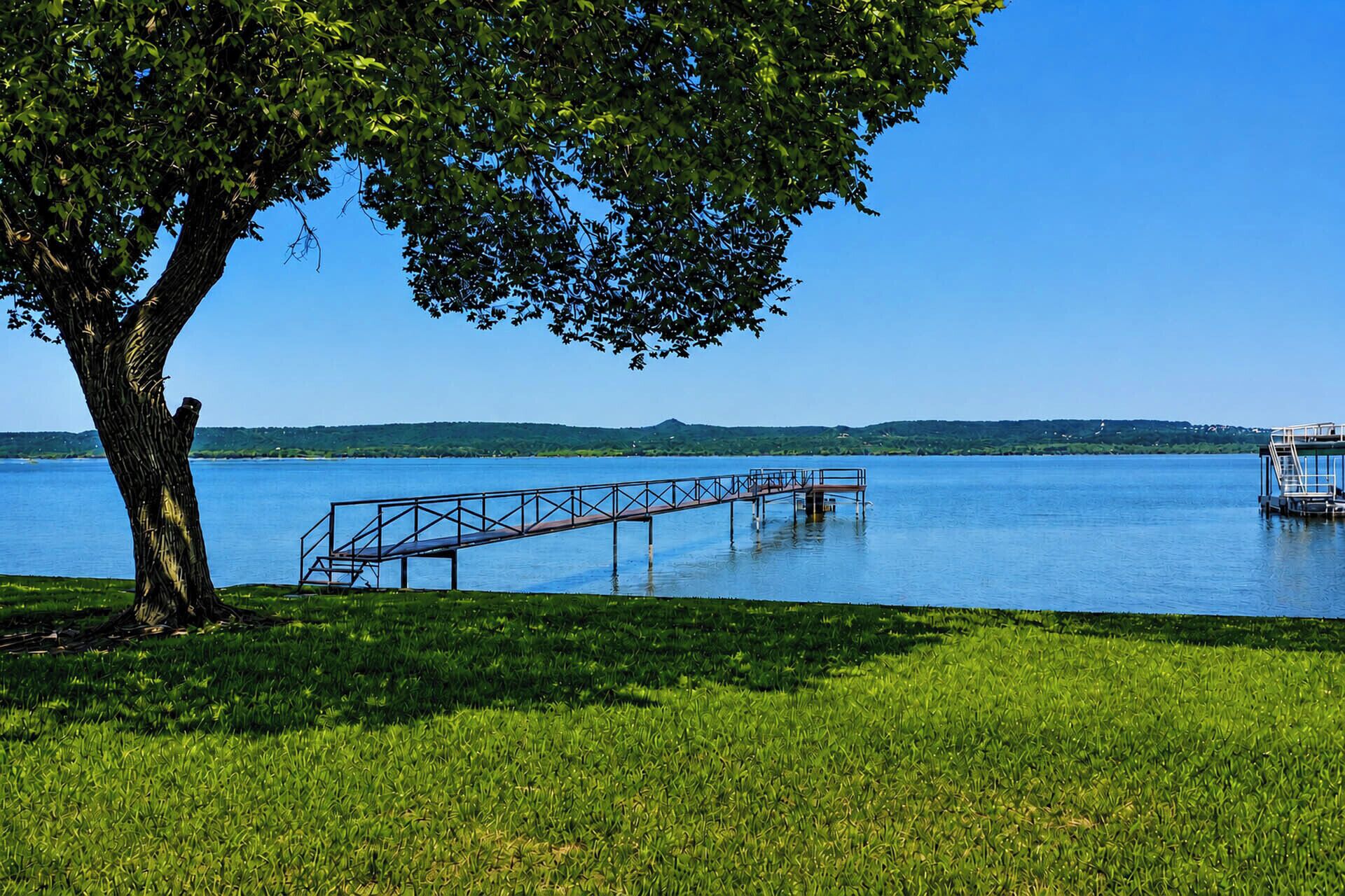A clear lake with a dock extending into the water, surrounded by grassy areas and a large tree providing shade.