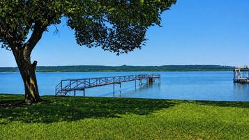 A clear lake with a dock extending into the water, surrounded by grassy areas and a large tree providing shade.