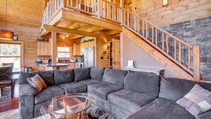 A spacious living area with a large gray sectional sofa, a glass coffee table, and a fireplace. A wooden staircase with a railing leads to an upper level. - Cozy Family Cabin: Game Room, Near National Parks! (Duck Creek Village)