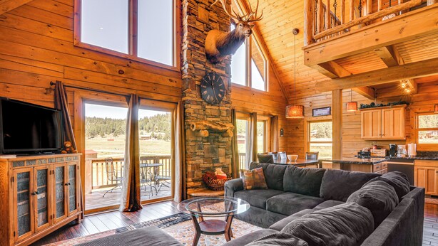 A log cabin interior with a stone fireplace, a mounted elk head, a large sofa, and a television on a wooden cabinet. - Cozy Family Cabin: Game Room, Near National Parks! (Duck Creek Village)