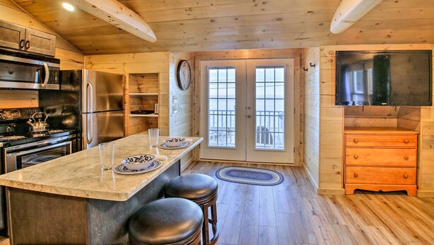 A kitchen with wooden walls, stainless steel appliances, a large island with stools, and a view through a glass door. - Sebago Lake Haven: Water Views, Steps to Shoreline (Sebago)