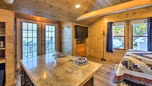 A wooden cabin interior with a kitchen area featuring a granite countertop, two windows with curtains, and a bed with a quilt. - Sebago Lake Haven: Water Views, Steps to Shoreline (Sebago)