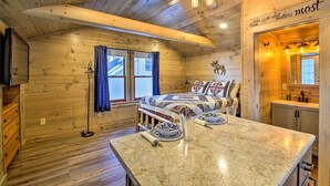 A cozy cabin interior with a wooden ceiling, a bed with patterned bedding, a dining table with plates and a glass, and a kitchen area. - Sebago Lake Haven: Water Views, Steps to Shoreline (Sebago)