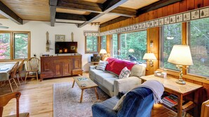 A cozy living room with a wooden ceiling, a sofa, a coffee table, and a TV. There are large windows with a view of trees. - Community Perks: Retreat w/ Thirteenth Lake Access (North River)