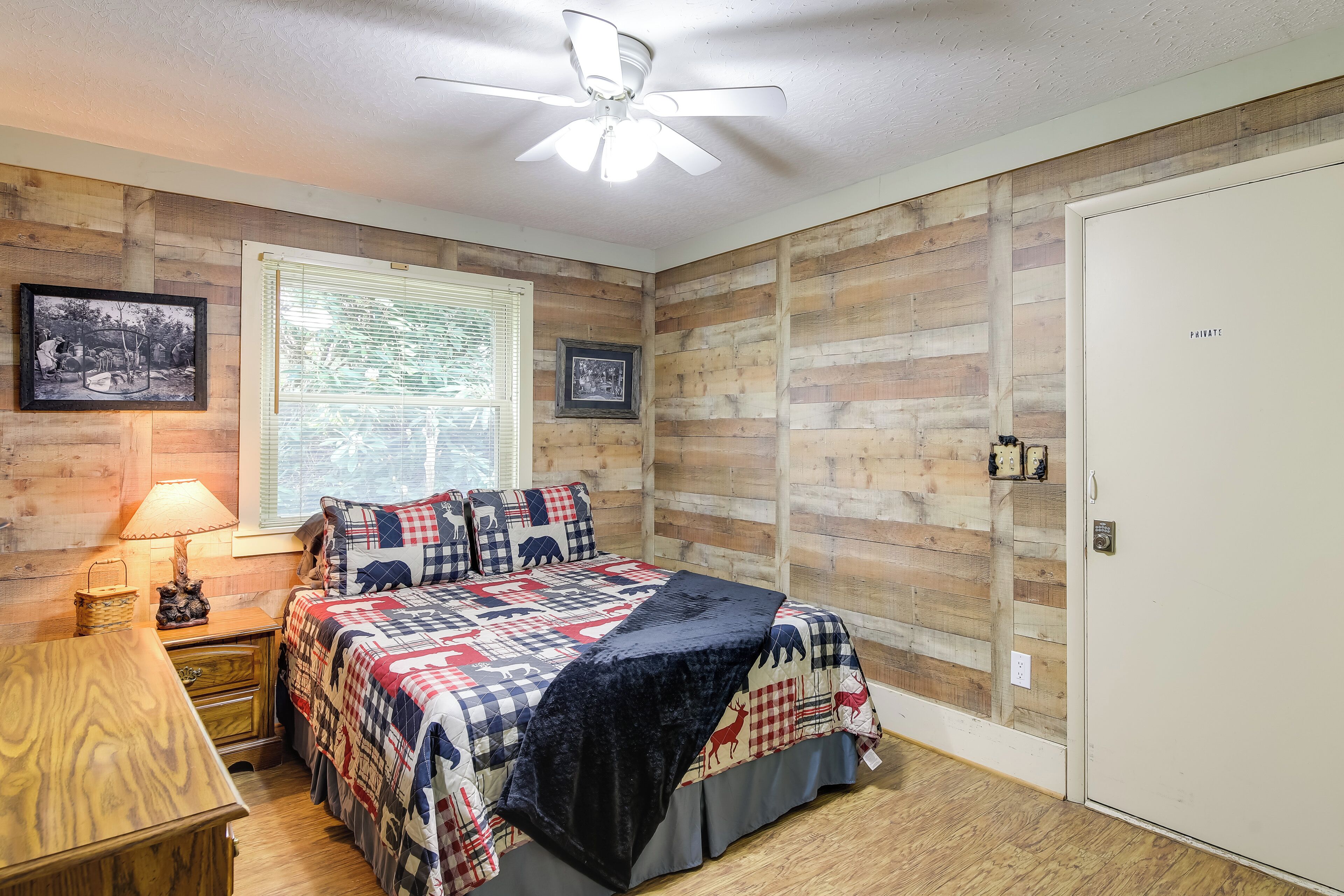 Ein Schlafzimmer mit einer Holzwand, einem Bett mit karierter Steppdecke, einem Deckenventilator und einem Fenster mit Jalousien.