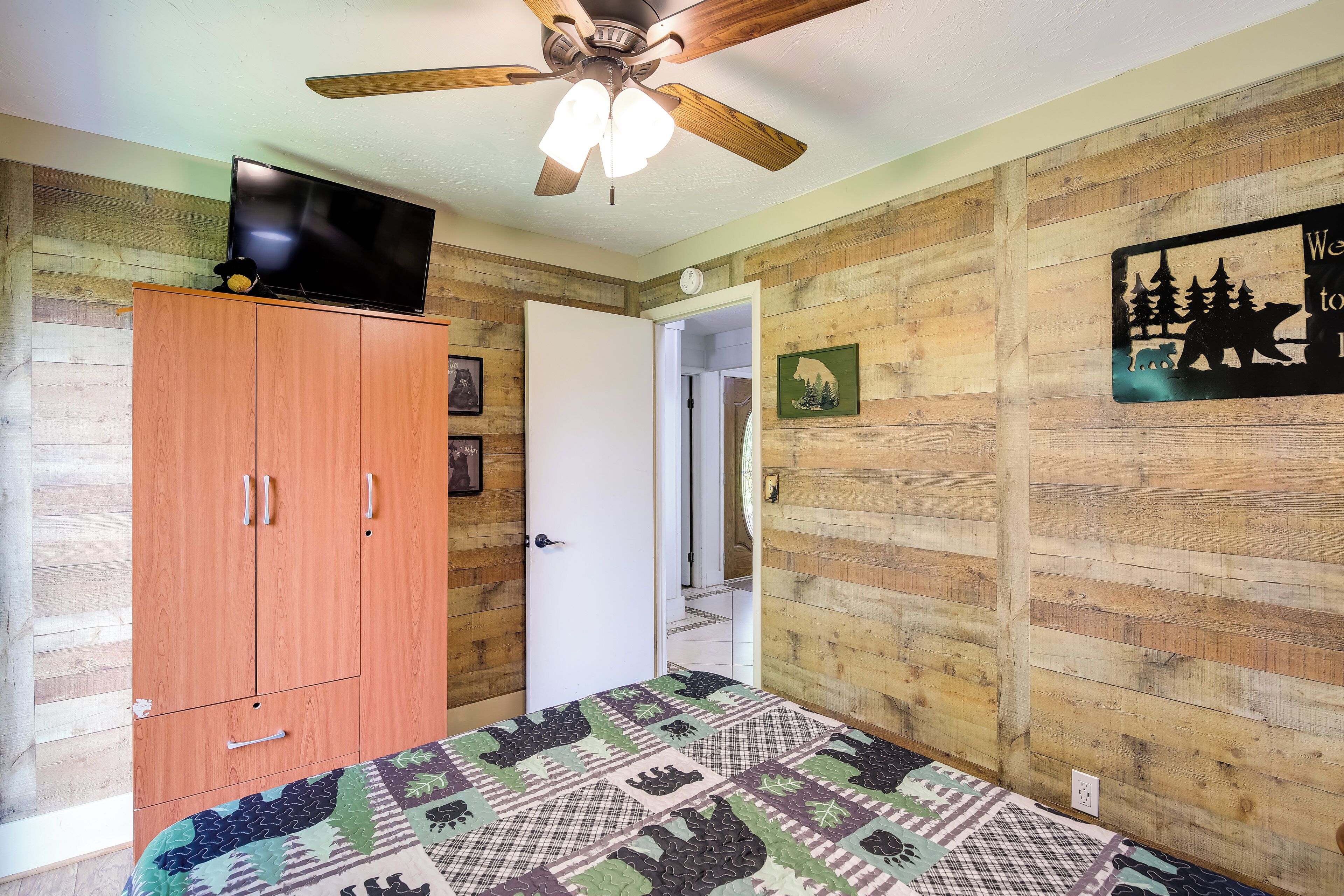Un dormitorio con pared de madera, ventilador de techo, cama con colcha estampada, televisor sobre un mueble de madera y puerta que conduce a otra habitación.