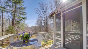 Eine Holzdielen-Terrasse mit Glastür zu einem überdachten Bereich, umgeben von Bäumen und blauem Himmel.