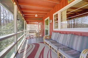 A covered porch with a sofa, chairs, and a railing overlooking a forested area.