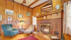 Ein gemütliches Wohnzimmer mit Holzwänden, einem Kamin aus Backsteinen, zwei Sesseln, einem roten Teppich und gerahmten Bildern an der Wand.
