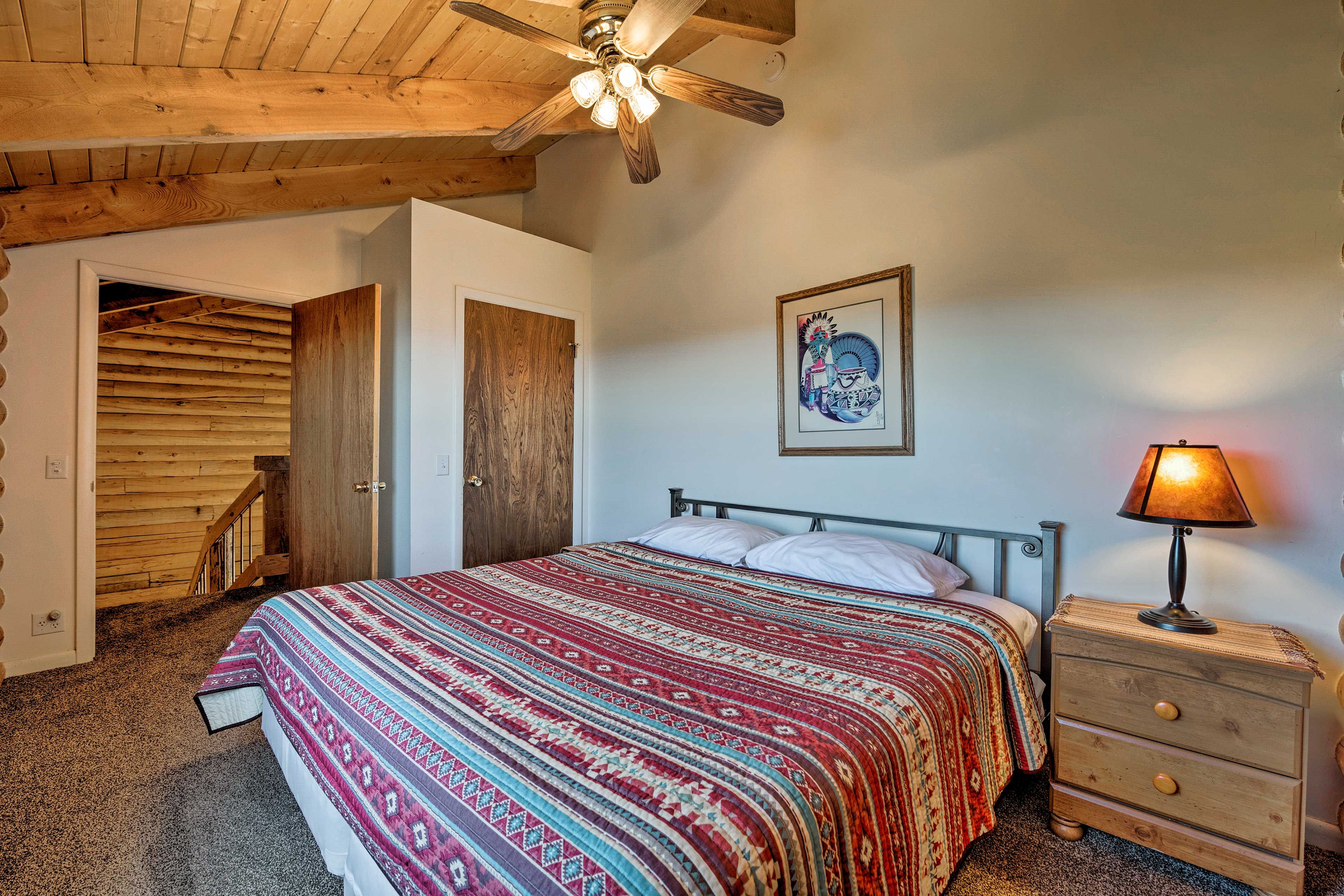 Un dormitorio con techo de madera, una cama con manta estampada, una mesita de noche con lámpara y una puerta que da a otra habitación.