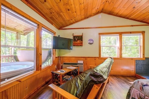 A cabin interior with a wooden ceiling, a large window, a television mounted on the wall, and a sofa with a cushion. - Secluded Nantahala 'gone Hunting' Cabin w/ Hot Tub (Bryson City)