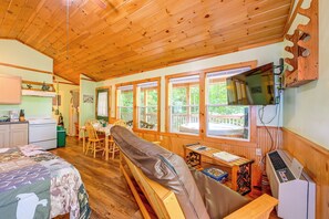 A cozy cabin interior with wooden walls and ceiling, a dining area with a table and chairs, a bed with a patterned blanket, and a television mounted on the wall.