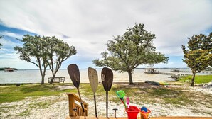 A wooden deck with beach gear, including paddles and a bucket, overlooking a beach with trees and a pier. - Waterfront Perdido Beach House w/ Canoes & Kayaks! (Elberta)