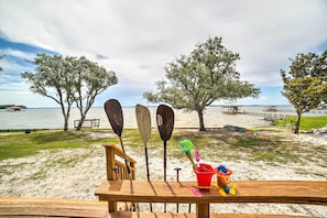 A wooden deck with beach gear, including paddles and a bucket, overlooking a beach with trees and a pier. - Waterfront Perdido Beach House w/ Canoes & Kayaks! (Elberta)