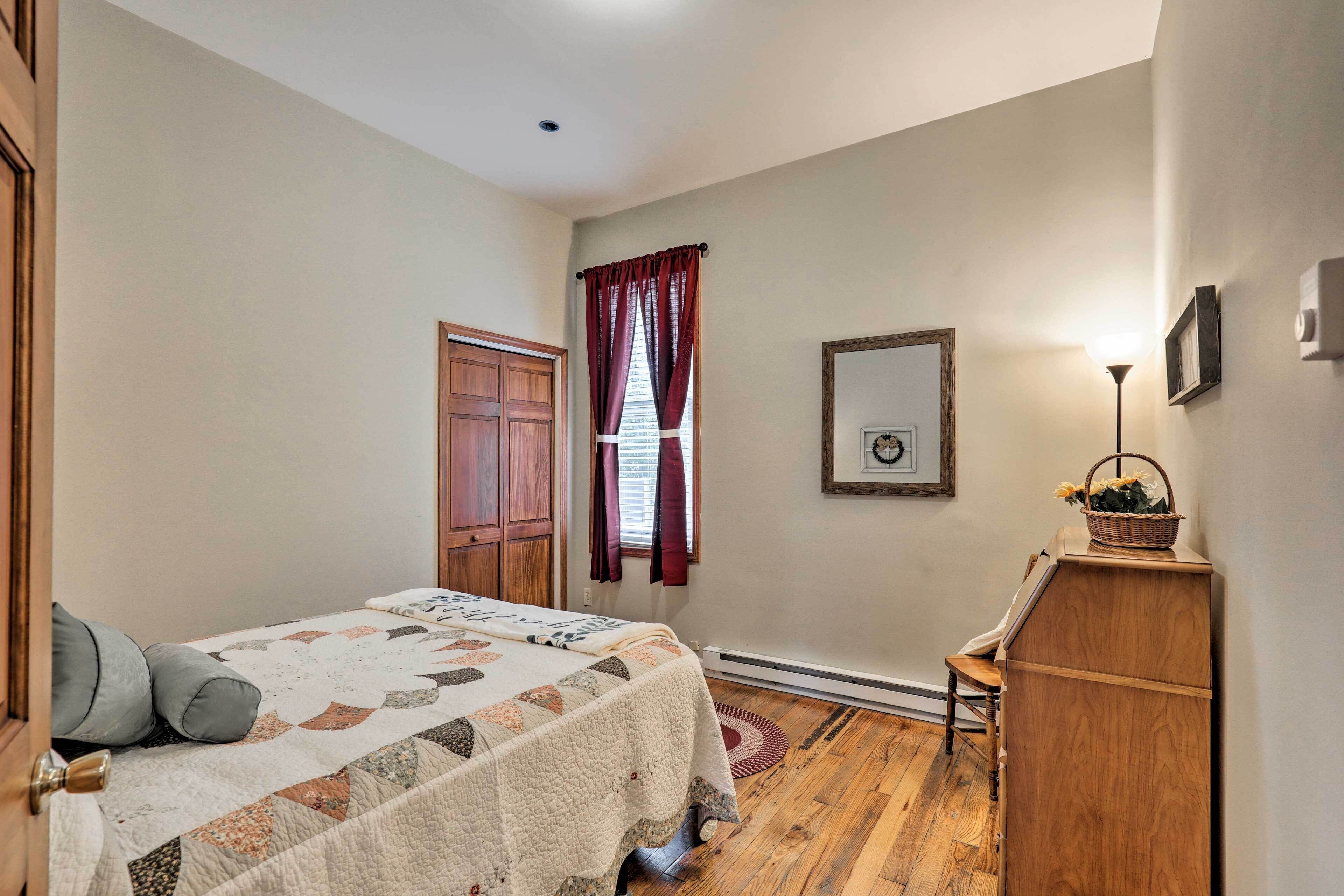 A bedroom with a bed, wooden wardrobe, a desk, a chair, a window with curtains, and a framed picture on the wall.