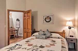 Un dormitorio con cama, una mesita de noche con lámpara, una puerta de madera, un cuadro enmarco y una pequeña mesa con sillas.