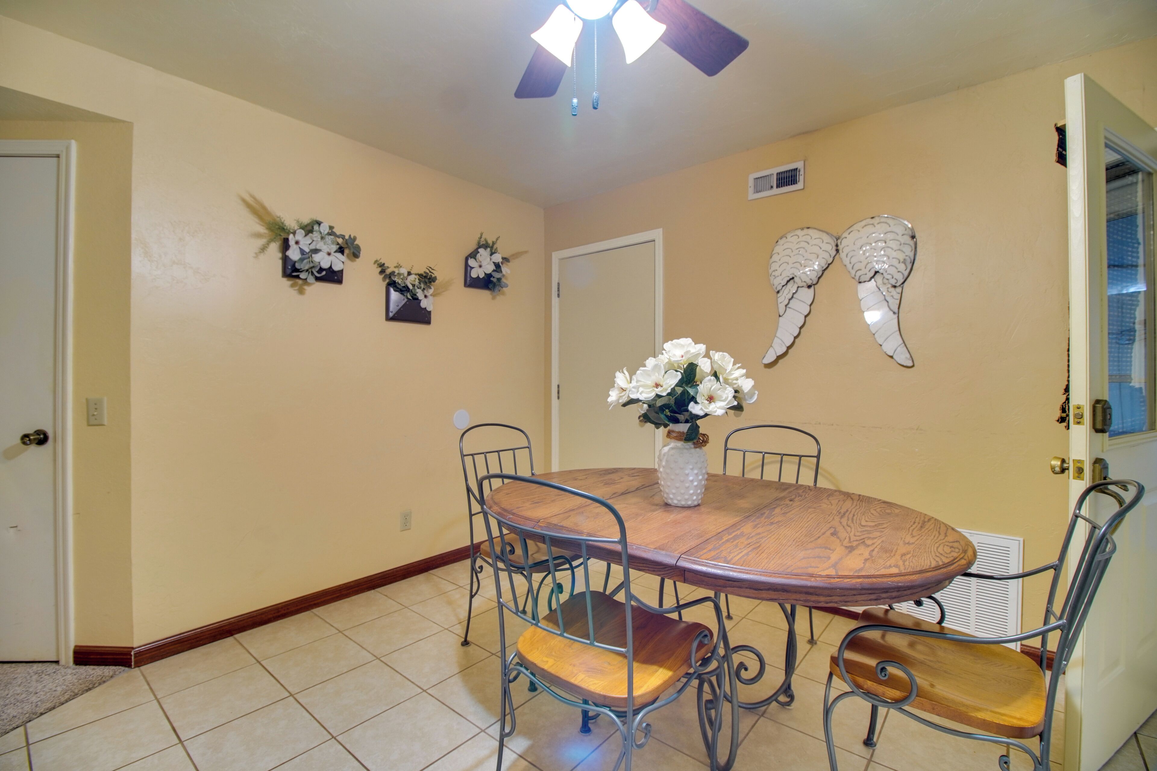 A dining area with a round wooden table, four metal chairs with cushioned seats, a vase with flowers, and decorative wall art.