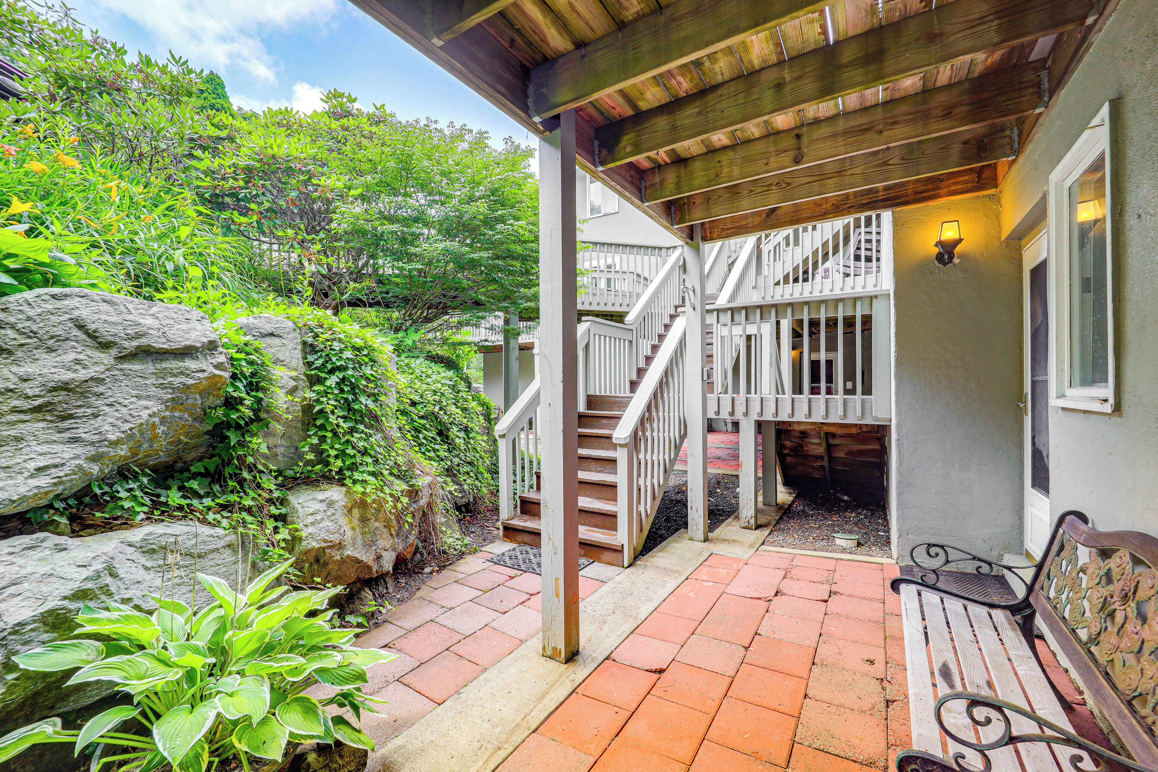 A covered outdoor area with a wooden staircase leading to a second floor, surrounded by greenery and a stone wall.