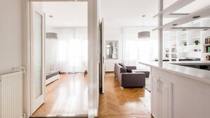 A modern living space with a wooden floor, a grey sofa, a kitchen area with white cabinetry, and a dining table. - Basilica View Apartment With Balcony (Budapest)