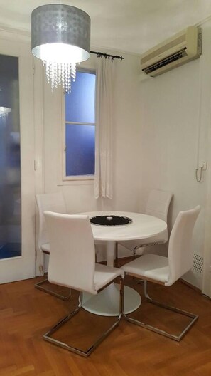 Un comedor con una mesa redonda, cuatro sillas, una lámpara de araña, una ventana con cortinas y un equipo de aire acondicionado.