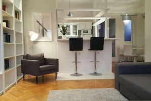 Una sala de estar moderna con un sofá gris, una estantería, dos taburetes negros y una alfombra pequeña.