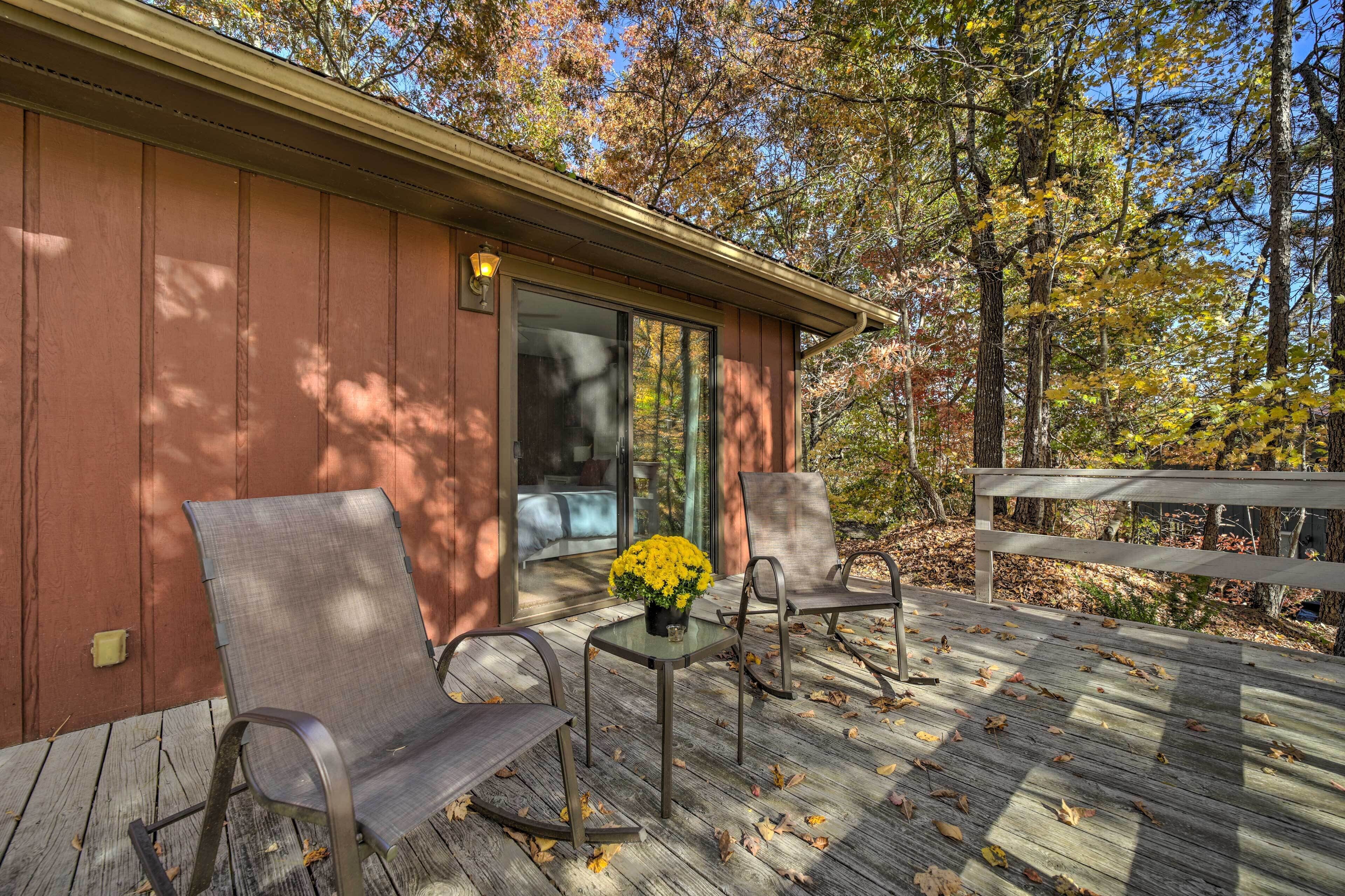A wooden deck with two chairs and a table, a glass door leading to a room, and a view of trees.