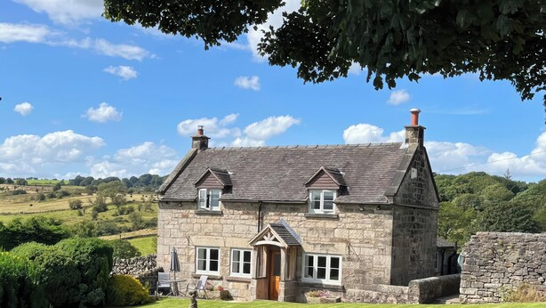 A stone house with a sloped roof, a front entrance, and a stone wall in the foreground. - Sycamore Cottage (Ashbourne)