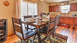 A dining area with a wooden table set for four, surrounded by chairs. A kitchen with wooden cabinets and a window is visible in the background. - Drake Home on Big Thompson River - 3 Mi. to Estes! (Drake)