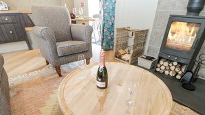 A cozy living room with a round wooden coffee table, a bottle of wine, and two glasses. A fireplace with logs is visible in the background. - Bolton Cottage (Appleby-in-Westmorland)