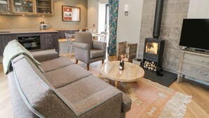 A cozy living room with a sofa, a coffee table, and a TV. There's a kitchen area with cabinets and a stove. - Bolton Cottage (Appleby-in-Westmorland)