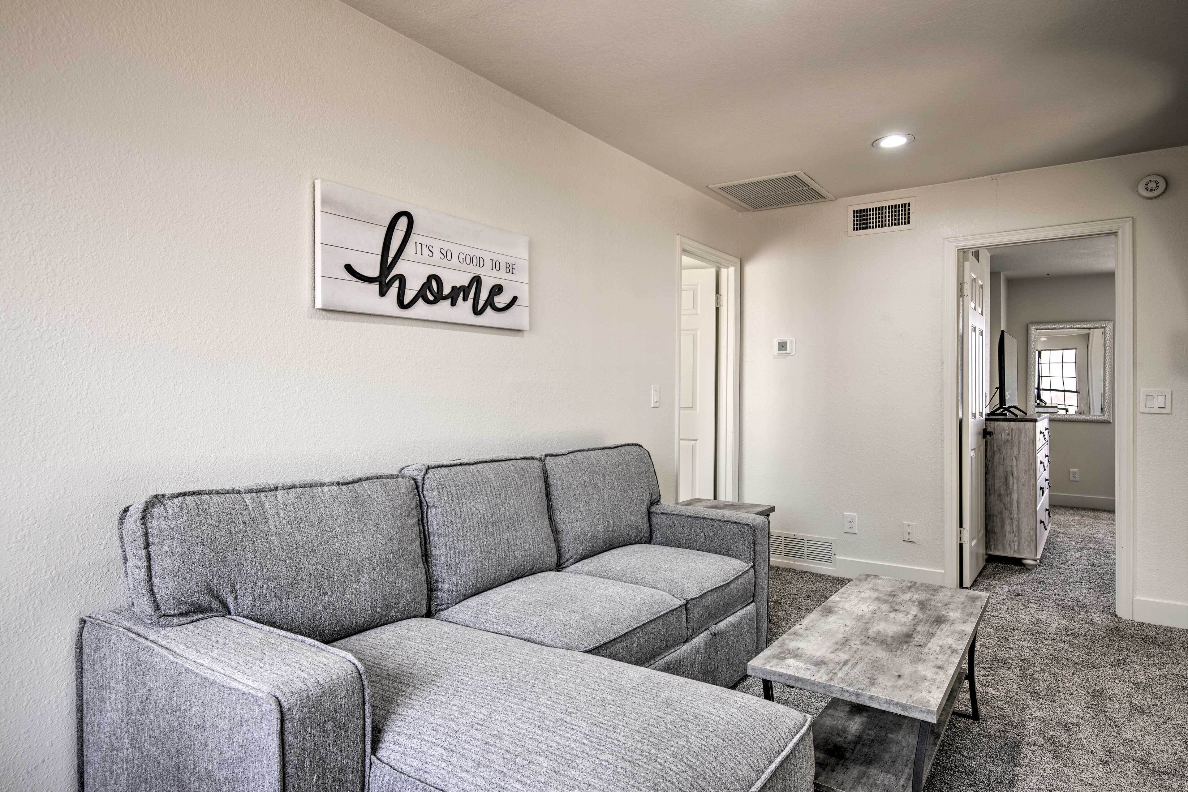 A living room with a gray sofa, a small coffee table, and a wall sign that says "It's so good to be home."