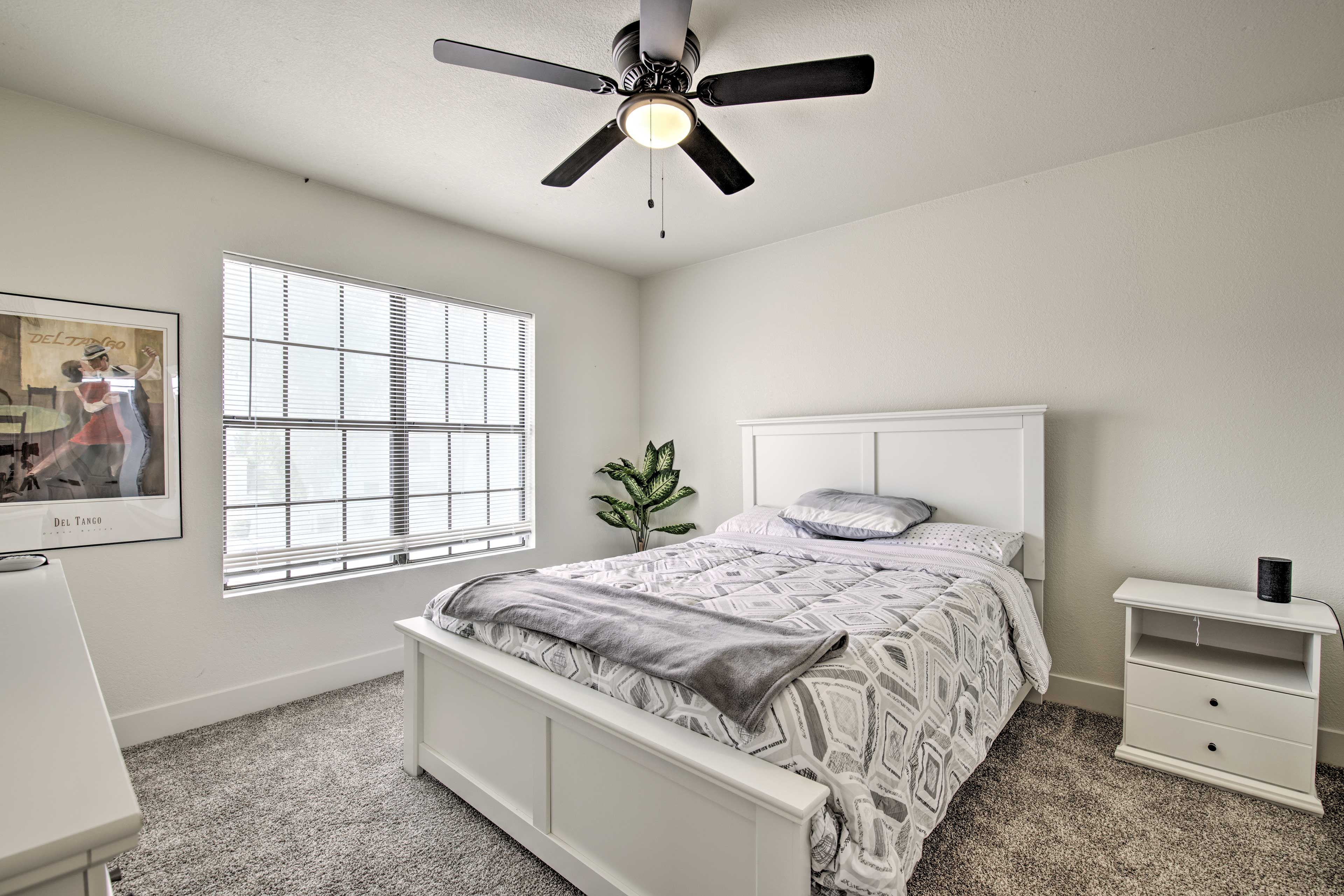 A bedroom with a bed, a nightstand, a ceiling fan, a window with blinds, and a framed picture on the wall.