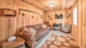 A cozy cabin interior with a wooden floor, a sofa, a chair, a fireplace, and wall decorations including a saw blade and antlers. - Remote Kentucky Cabin w/ Fire Pit & 2 Ponds (New Concord)