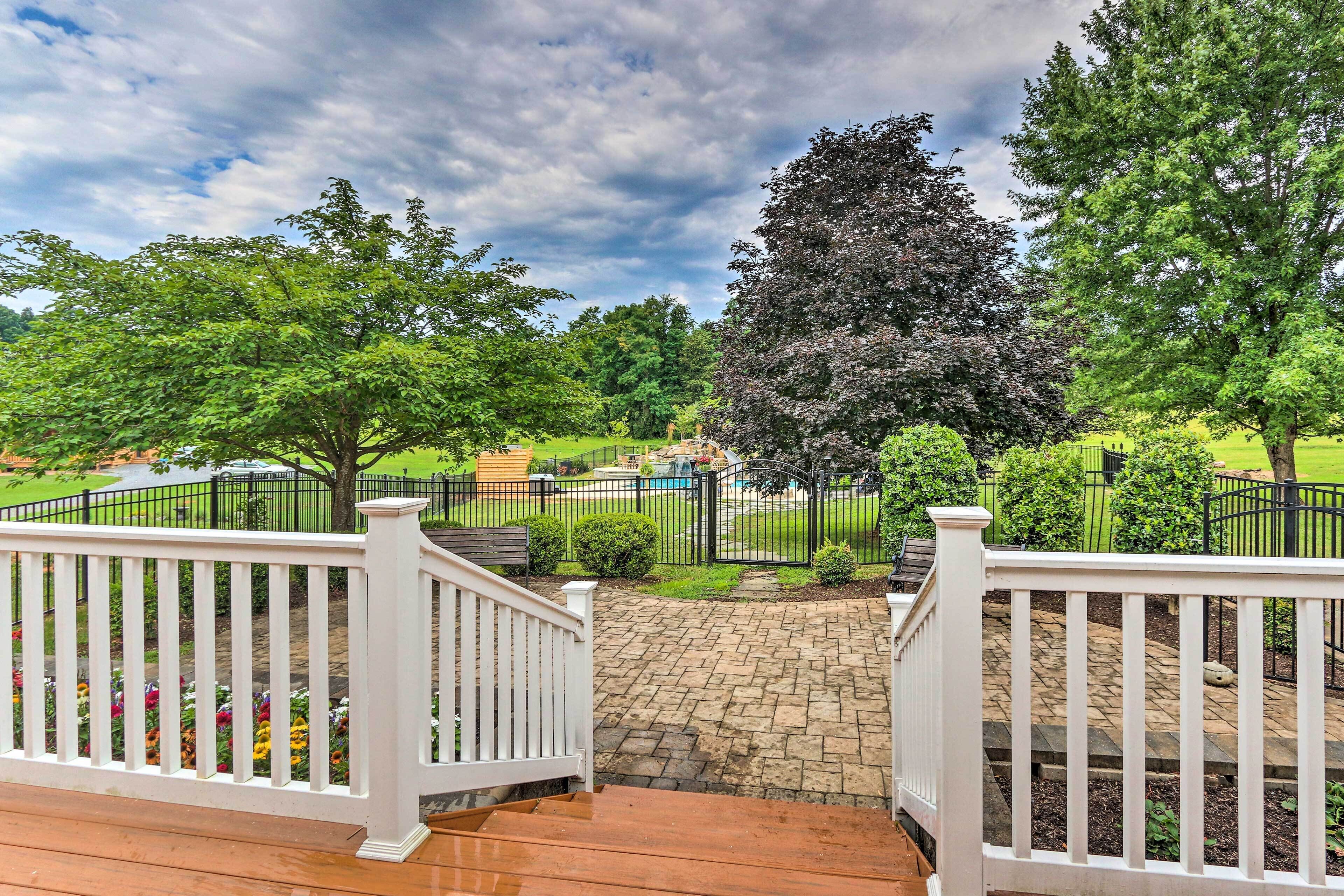 A wooden deck with white railings, a stone pathway, and a view of a well-maintained garden with various trees and a swimming pool.
